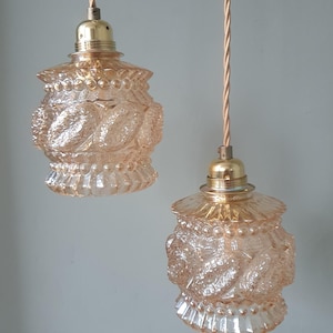 Op de afbeelding: Twee vintage hanglampen met perzikroze glazen kappen. De kappen hebben een gestructureerd, sierlijk ontwerp met een kralen boven- en onderkant. De lampen hangen aan goudkleurige armaturen en gedraaide snoeren.