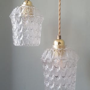 Op de afbeelding: Twee heldere glazen hanglampen met een textuurpatroon en goudkleurige hardware. De lampen hangen aan gedraaide beige snoeren. De glazen kappen hebben een uniek, getextureerd ontwerp en geven de armaturen een vintage uitstraling.
