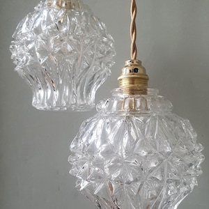 Könnte beinhalten: Zwei transparente Glas-Pendelleuchten mit einem rautenförmigen Muster. Die Leuchten haben goldfarbene Beschläge und gedrehte Kabel. Die Lampen hängen vor einem hellgrauen Hintergrund.