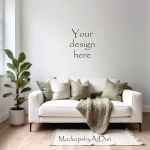 Puede incluir: Un sofá blanco con patas negras está adornado con cojines verdes y crema y una manta de punto grueso. Una planta en maceta está a la izquierda, y la pared sobre el sofá dice "Your design here."