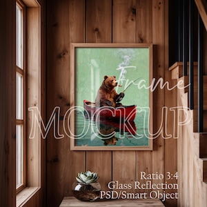 May include: A framed art print featuring a bear in a red canoe, smoking a pipe while fishing. The artwork has a green and blue background with a reflection of the bear and canoe in the water. The frame is a light brown color.