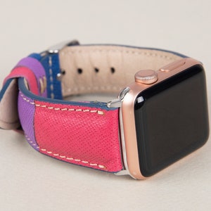 Pode incluir: Uma banda de Apple Watch de couro rosa, azul e roxo com costuras brancas. A banda está presa a um Apple Watch de ouro rosa.