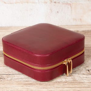 May include: A square, burgundy jewelry box with a gold-colored zipper. The box is made of a smooth material and sits on a wooden surface. The zipper has two pulls.