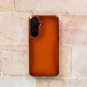 May include: A brown leather phone case with a triple-lens camera cutout. The case has a smooth, polished finish and a rectangular shape. The phone case is shown against a textured, light-colored brick wall.