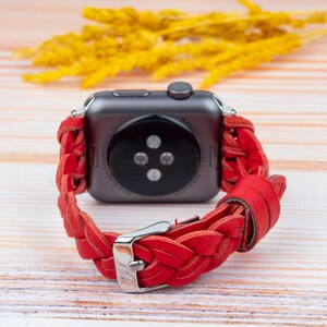 Puede incluir: Pulsera de reloj de cuero trenzado rojo para un Apple Watch. La pulsera est&aacute; tejida con un patr&oacute;n trenzado y tiene una hebilla plateada.