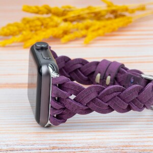 Pu&ograve; includere: Un cinturino per orologio in pelle intrecciata viola per un Apple Watch. Il cinturino &egrave; realizzato in pelle morbida e flessibile e presenta un design intrecciato. Il cinturino &egrave; mostrato su una superficie in legno chiaro.