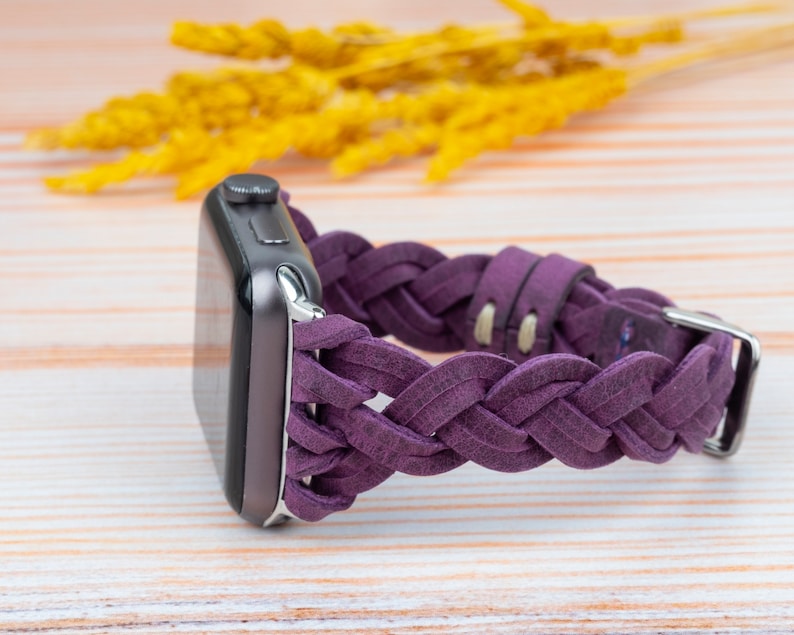 Pu&ograve; includere: Un cinturino per orologio in pelle intrecciata viola per un Apple Watch. Il cinturino &egrave; realizzato in pelle morbida e flessibile e presenta un design intrecciato. Il cinturino &egrave; mostrato su una superficie in legno chiaro.