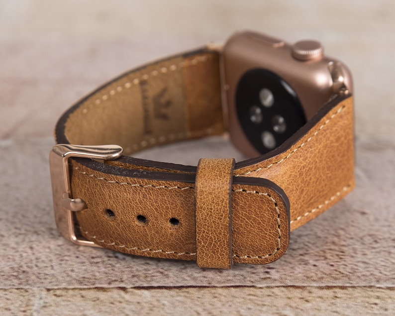 May include: A brown leather watch strap with a rose gold buckle and a stitched edge. The strap is designed for an Apple Watch.
