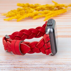 Puede incluir: Una correa de reloj de cuero trenzado roja para un Apple Watch. La correa est&aacute; hecha de cuero trenzado y tiene una hebilla plateada.