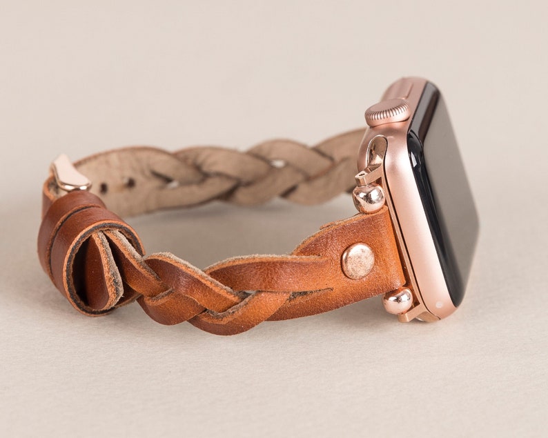 Puede incluir: Una correa de reloj trenzada de cuero marr&oacute;n con un Apple Watch de oro rosa.