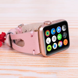 Peut inclure: Une Apple Watch en or rose avec un bracelet en cuir rose. Le cadran de la montre affiche une vari&eacute;t&eacute; d'ic&ocirc;nes d'applications, notamment une horloge, une boussole et un moniteur de fr&eacute;quence cardiaque.