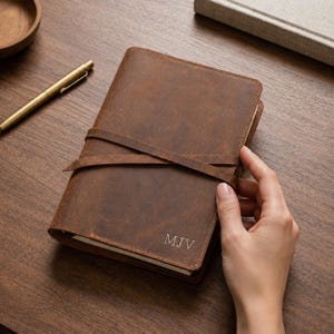 May include: A brown leather journal with a strap closure and the initials "MJV" embossed on the cover. The journal is open, revealing the pages inside. A gold pen and wooden bowl are visible on the wooden surface.