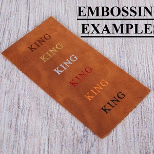 Pu&ograve; includere: Un pezzo rettangolare di pelle marrone con il testo EMBOSSING EXAMPLES in alto. Sotto, la parola KING &egrave; impressa sei volte in una colonna diagonale, visualizzata in diversi colori tra cui oro, argento, rosso, arancione e nero.