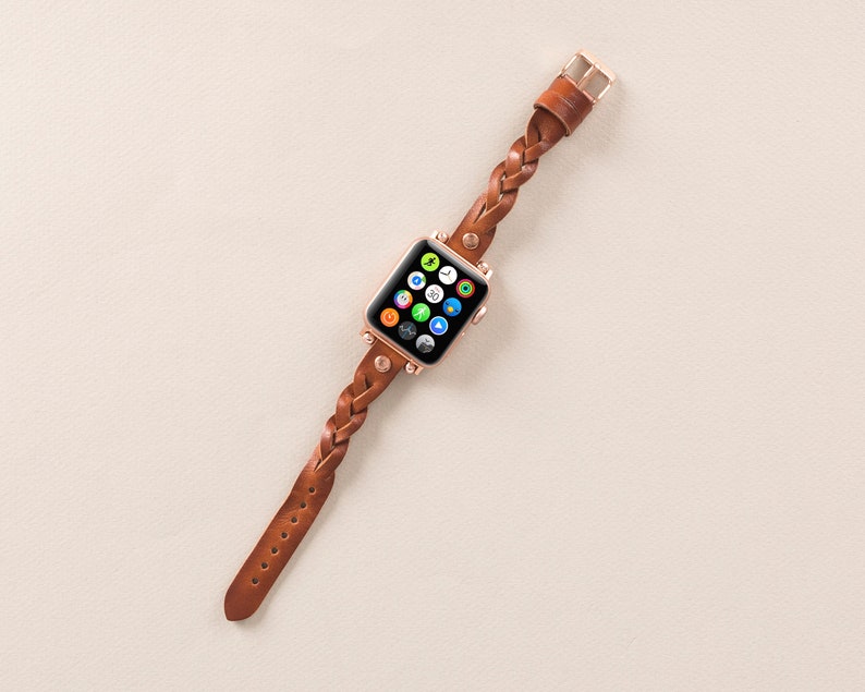 Puede incluir: Una correa de reloj trenzada de cuero marr&oacute;n con un Apple Watch de oro rosa con una esfera blanca y coloridos iconos de aplicaciones.