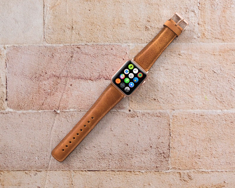 May include: A brown leather watch strap for an Apple Watch. The strap is shown against a brick wall background.