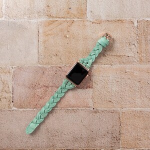 May include: A mint green braided leather watch band with a rose gold buckle and a black smartwatch face. The watch band is made of a soft, flexible leather and is designed to fit a variety of wrist sizes.