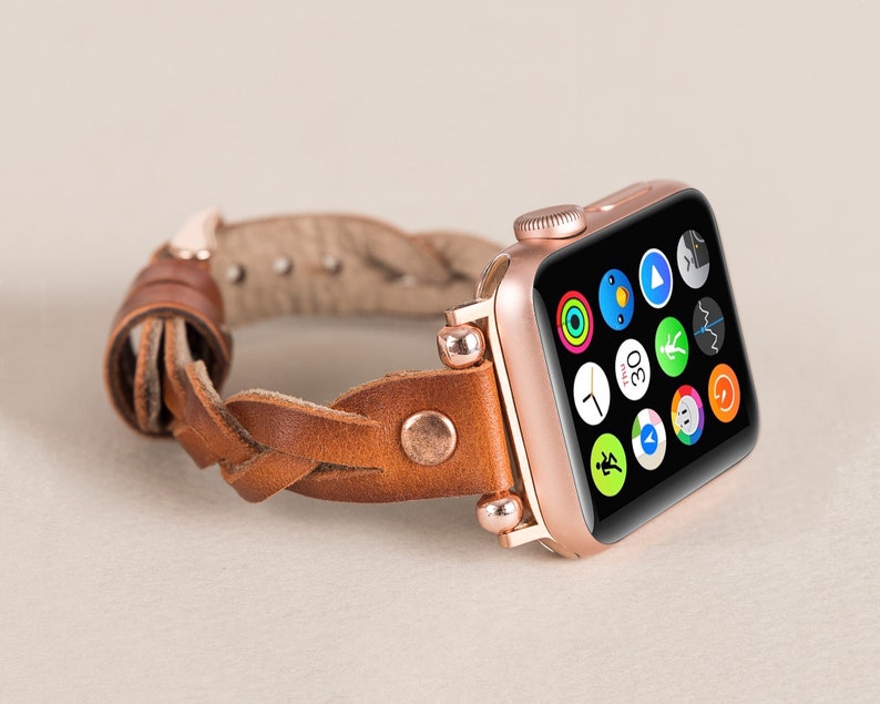 Puede incluir: Un Apple Watch de oro rosa con una correa de cuero marr&oacute;n trenzada. La esfera del reloj muestra una variedad de iconos de aplicaciones.