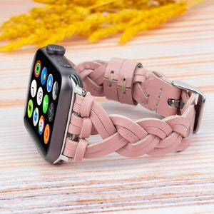 K&ouml;nnte beinhalten: Ein rosafarbenes geflochtenes Lederarmband f&uuml;r eine Apple Watch. Das Armband ist aus weichem, geschmeidigem Leder gefertigt und hat ein geflochtenes Design. Das Armband wird auf einer hellen Holzoberfl&auml;che gezeigt.