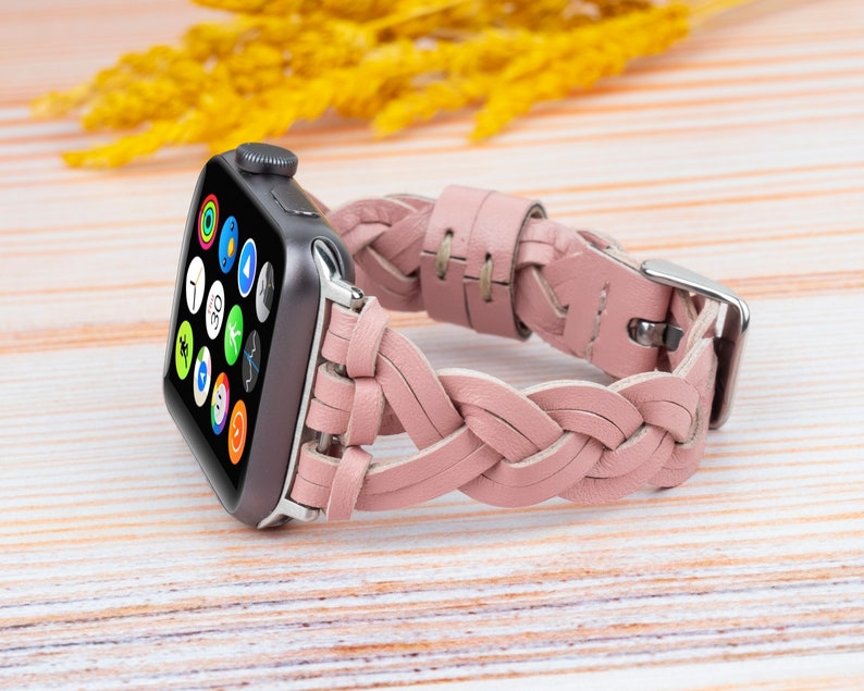 K&ouml;nnte beinhalten: Ein rosafarbenes geflochtenes Lederarmband f&uuml;r eine Apple Watch. Das Armband ist aus weichem, geschmeidigem Leder gefertigt und hat ein geflochtenes Design. Das Armband wird auf einer hellen Holzoberfl&auml;che gezeigt.