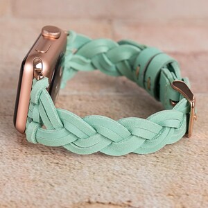 May include: A rose gold Apple Watch with a mint green braided leather band. The band features a woven design and a gold-colored buckle. The watch is displayed on a light brown surface.
