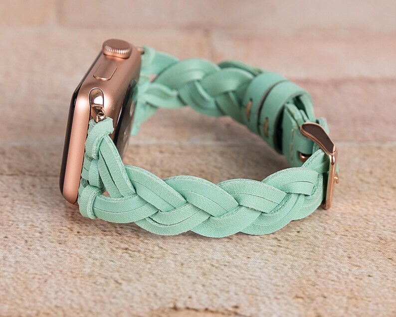 May include: A rose gold Apple Watch with a mint green braided leather band. The band features a woven design and a gold-colored buckle. The watch is displayed on a light brown surface.