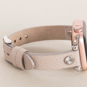 Pode incluir: Uma pulseira de rel&oacute;gio de couro cinza claro com um Apple Watch em ouro rosa. A pulseira tem um &uacute;nico strass no lado.