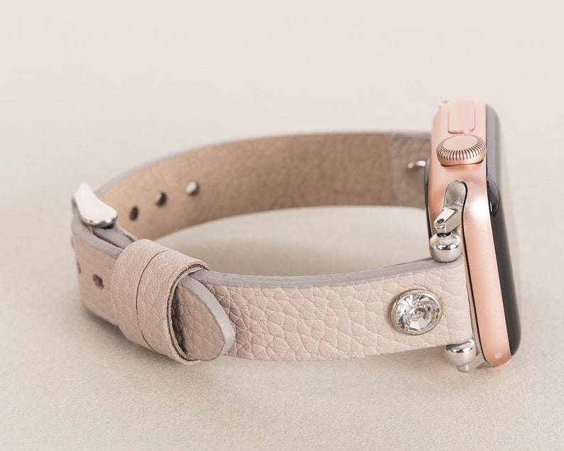 Pode incluir: Uma pulseira de rel&oacute;gio de couro cinza claro com um Apple Watch em ouro rosa. A pulseira tem um &uacute;nico strass no lado.