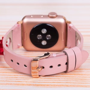 Peut inclure: Un bracelet de montre en cuir rose avec une boucle en or rose et une Apple Watch en or rose.