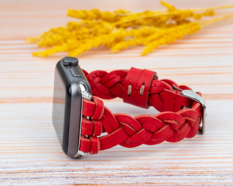 Puede incluir: Una correa de reloj de cuero trenzado roja con una hebilla plateada. La correa est&aacute; dise&ntilde;ada para un Apple Watch.