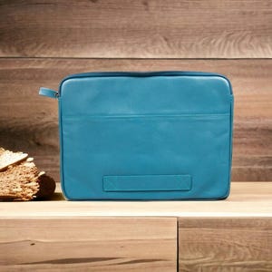 May include: A teal-colored rectangular laptop sleeve with a zipper closure and a handle. The sleeve is made of a smooth material and has a stitched design. The background is a wooden surface.