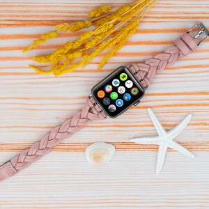 K&ouml;nnte beinhalten: Ein rosafarbenes geflochtenes Lederarmband mit einem silbernen Apple Watch-Zifferblatt, auf dem verschiedene App-Symbole angezeigt werden. Die Uhr liegt auf einem hellen Holzhintergrund mit einem wei&szlig;en Seestern und einer Muschel.