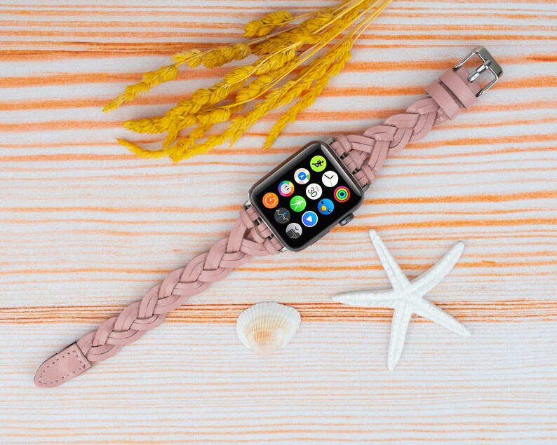 K&ouml;nnte beinhalten: Ein rosafarbenes geflochtenes Lederarmband mit einem silbernen Apple Watch-Zifferblatt, auf dem verschiedene App-Symbole angezeigt werden. Die Uhr liegt auf einem hellen Holzhintergrund mit einem wei&szlig;en Seestern und einer Muschel.