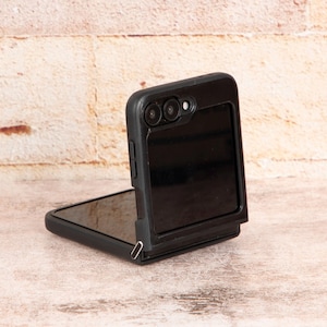 May include: A black foldable phone with a dual camera lens on the back. The phone is closed and the screen is facing away from the viewer.