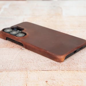 Puede incluir: Una funda de teléfono de cuero marrón con un recorte para la cámara. La funda tiene un aspecto liso y desgastado y un color marrón oscuro. La funda está diseñada para proteger el teléfono.