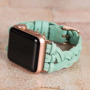 May include: A rose gold Apple Watch with a black screen and a braided mint green leather band. The band features a gold-colored buckle. The watch is displayed on a light brown surface.