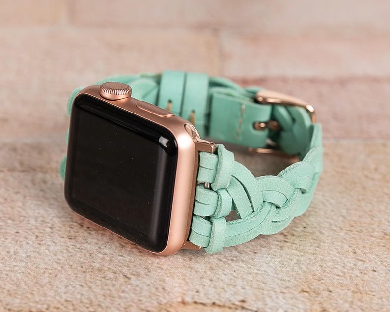 May include: A rose gold Apple Watch with a black screen and a braided mint green leather band. The band features a gold-colored buckle. The watch is displayed on a light brown surface.