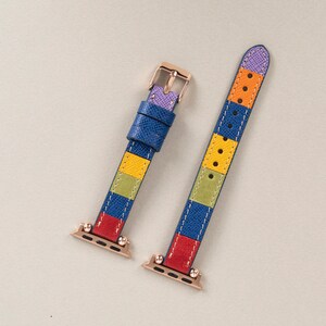 K&ouml;nnte beinhalten: Ein farbenfrohes Armband mit einem gestreiften Muster in Blau, Gelb, Gr&uuml;n, Rot und Lila. Das Armband ist aus Leder und hat eine ros&eacute;goldene Schlie&szlig;e.