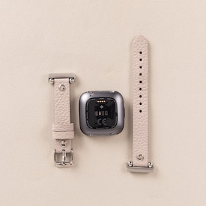 Puede incluir: Una correa de reloj de cuero gris claro con una hebilla plateada y un rastreador de fitness gris con una pantalla negra y el texto "CE" en ella.