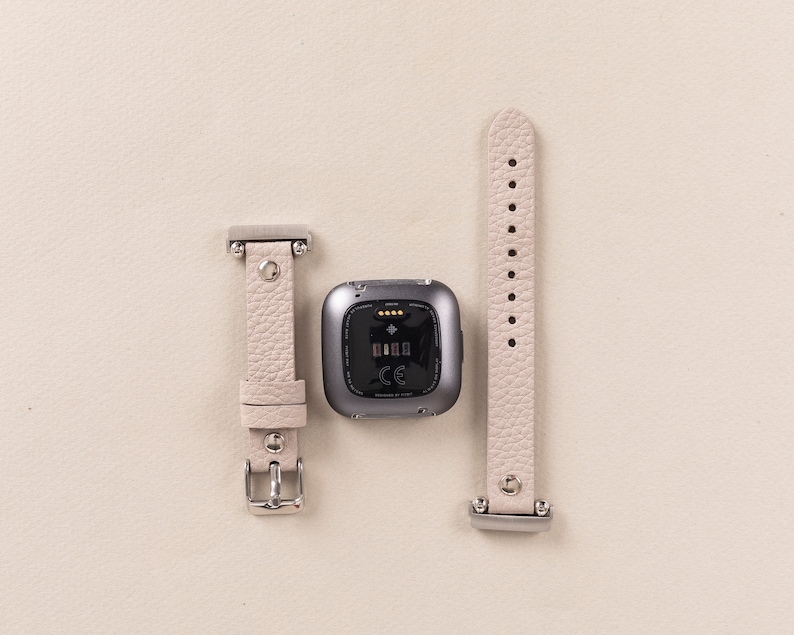Puede incluir: Una correa de reloj de cuero gris claro con una hebilla plateada y un rastreador de fitness gris con una pantalla negra y el texto "CE" en ella.