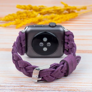 Pu&ograve; includere: Un cinturino in pelle intrecciata viola per uno smartwatch. Il cinturino &egrave; realizzato in pelle morbida e flessibile e presenta un design intrecciato. Il cinturino &egrave; mostrato su una superficie in legno.