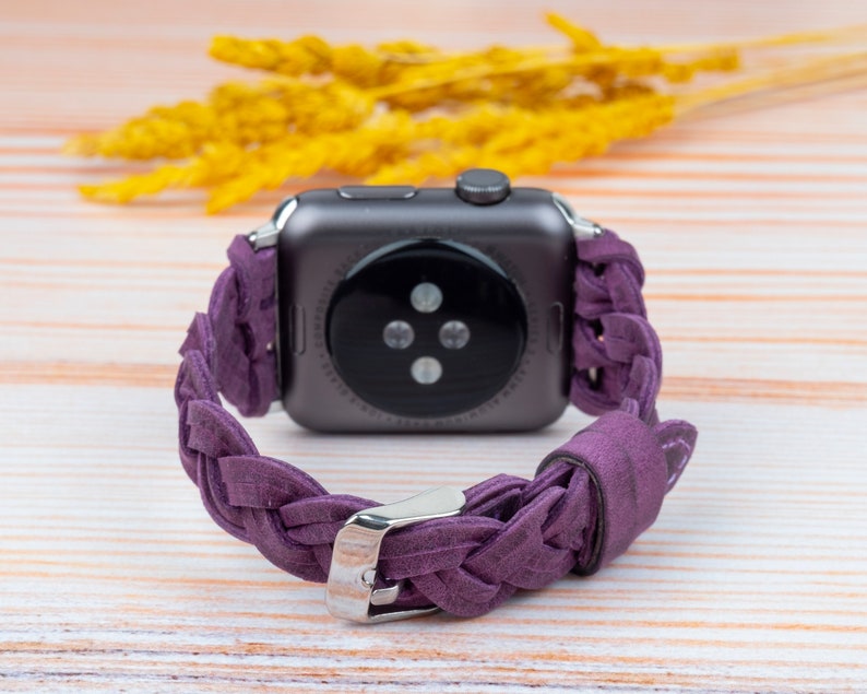 Pu&ograve; includere: Un cinturino in pelle intrecciata viola per uno smartwatch. Il cinturino &egrave; realizzato in pelle morbida e flessibile e presenta un design intrecciato. Il cinturino &egrave; mostrato su una superficie in legno.
