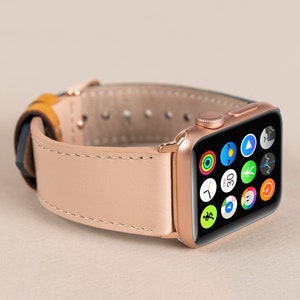 May include: A rose gold Apple Watch with a light pink leather strap. The watch face shows a range of app icons.