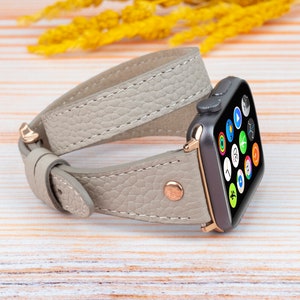 Puede incluir: Una correa de Apple Watch de cuero gris claro con un diseño de doble vuelta. La correa tiene una hebilla de oro rosa y se muestra sobre una superficie de madera.