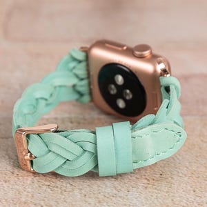 May include: A rose gold Apple Watch with a braided mint green leather band. The band features a gold-colored buckle and a braided design. The watch is displayed on a light brown surface.