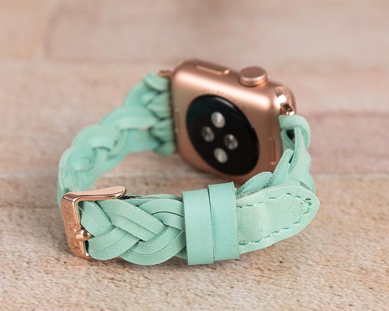 May include: A rose gold Apple Watch with a braided mint green leather band. The band features a gold-colored buckle and a braided design. The watch is displayed on a light brown surface.