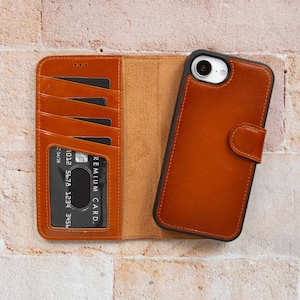 Könnte beinhalten: Eine braune Leder-Handyhülle mit Brieftaschen-Design. Die Hülle ist offen und zeigt Kartenfächer und eine Kreditkarte. Eine zweite Handyhülle ist geschlossen, mit einem Magnetverschluss. Auf einem Ziegelhintergrund.