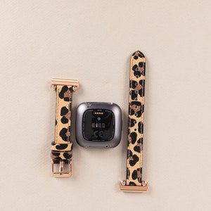 Puede incluir: Un rastreador de fitness negro con una banda de reloj de estampado de leopardo. La banda tiene detalles dorados. El rastreador muestra la hora y la fecha.