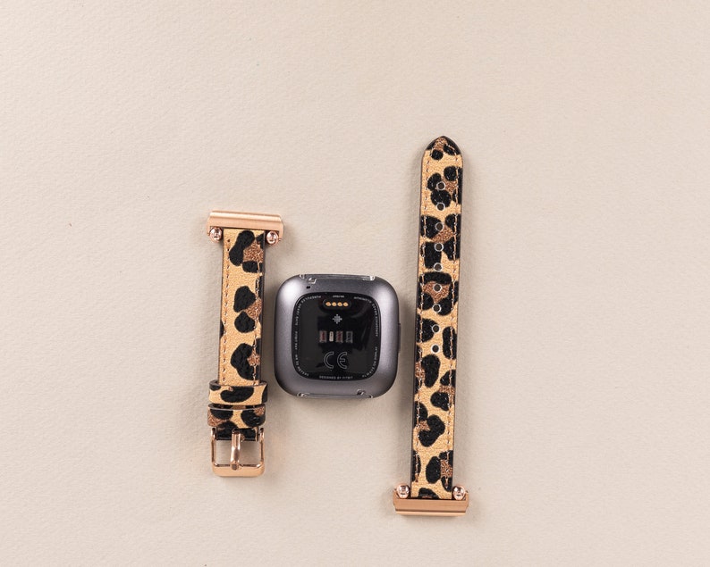 Puede incluir: Un rastreador de fitness negro con una banda de reloj de estampado de leopardo. La banda tiene detalles dorados. El rastreador muestra la hora y la fecha.