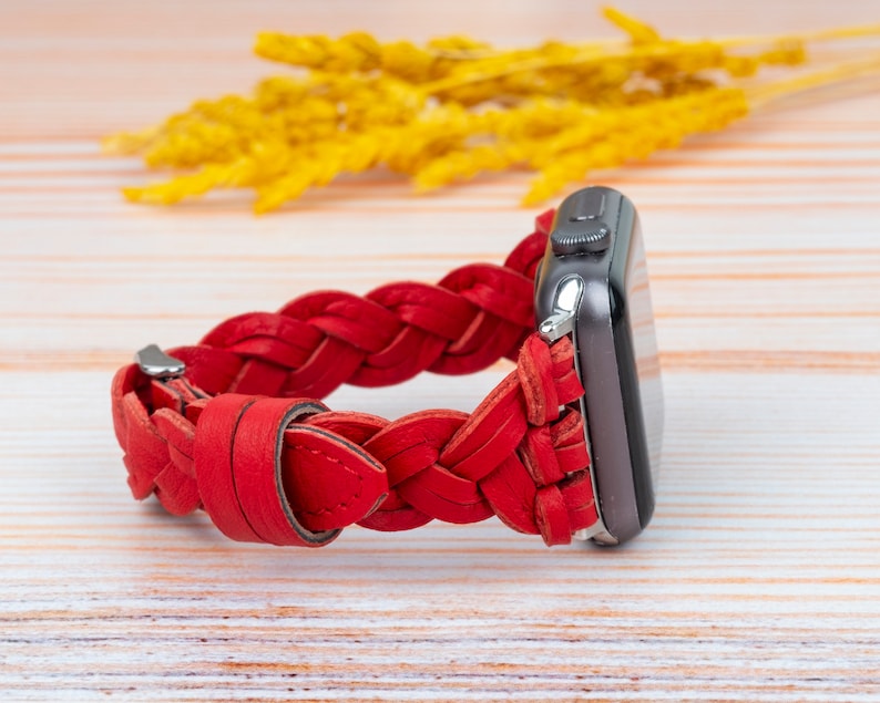 Puede incluir: Una correa de reloj de cuero trenzado roja para un Apple Watch. La correa est&aacute; hecha de cuero trenzado y tiene una hebilla plateada.