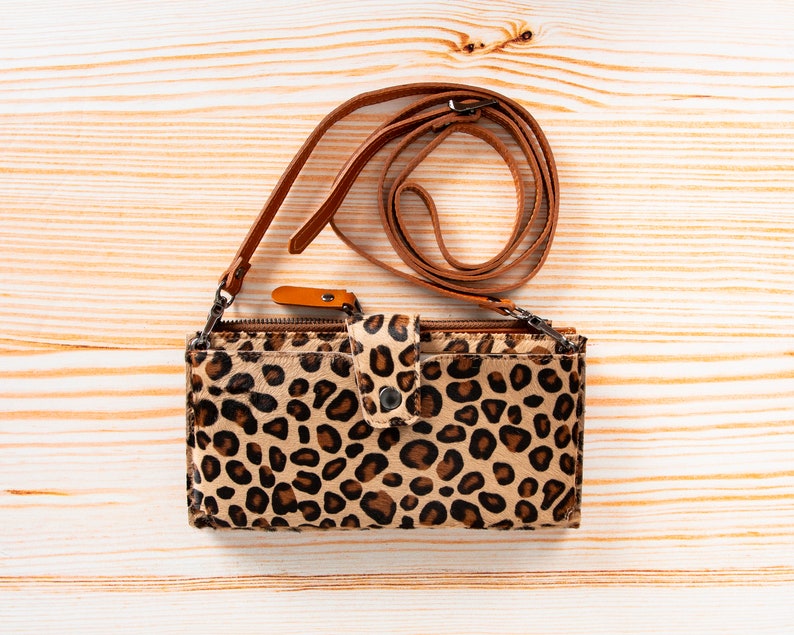 May include: A rectangular crossbody bag featuring a tan and black leopard print pattern. The bag includes a top zipper closure, a front flap with a metal snap button, and a thin, adjustable brown leather shoulder strap attached with metal clips.